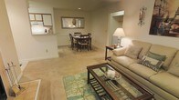 This single-floor space showcases an open-concept layout that connects the living and dining areas, creating a cohesive and functional environment. The living area is anchored by a plush, neutral-toned carpet layered with a light blue patterned area rug, providing a comfortable and inviting atmosphere. A glass-topped coffee table with a metal frame serves as a central accent piece, complemented by a cushioned sofa adorned with decorative pillows and a warm table lamp that adds a soft glow to the space.

The dining area flows naturally from the living space, featuring a dark wood dining table surrounded by matching chairs, ideal for everyday meals or casual gatherings. A framed mirror with chandelier detailing is mounted on the wall above, reflecting light and adding visual depth to the room. Recessed ceiling lighting illuminates the space efficiently throughout.

A partial view of the kitchen is visible through a pass-through counter, suggesting a convenient connection between cooking and living areas. An adjacent alcove or hallway leads to additional rooms. Soft, neutral wall tones and light carpeting throughout contribute to a calm and welcoming ambiance.