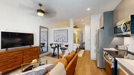 This thoughtfully designed single-story unit maximizes its compact footprint through an efficient open-concept layout. The living area serves as the heart of the space, featuring light wood flooring, recessed lighting, and ceiling fans that contribute to a bright, airy atmosphere. A comfortable seating arrangement anchors the living zone, complemented by a dining nook with bar-height seating and a dedicated media area. The kitchen, seamlessly integrated along one wall, is equipped with stainless steel appliances including a refrigerator, range, and dishwasher, set against sleek gray cabinetry.

The generously proportioned bedroom offers hardwood flooring, white walls, and a ceiling fan, creating a calm and restful environment. A closet and full-length mirror provide practical storage and functionality. Framed artwork and warm lighting contribute to a polished, cohesive aesthetic throughout.

The bathroom is finished with large-format neutral floor tiles, a glass-enclosed walk-in shower with tile surround, and a gray vanity, all accented with matte black hardware for a contemporary touch. Overall, the space presents a clean, modern design that balances style and practicality.