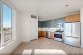 Located at 17 Avenue E in Bayonne, NJ, this thoughtfully designed one-bedroom, one-bathroom residence offers a clean, modern aesthetic throughout its well-utilized single-floor layout. Light oak hardwood flooring runs continuously through the space, complementing the crisp white walls and recessed lighting that give each room a bright, airy feel.

The open-concept kitchen and living area serves as the heart of the home. The kitchen is outfitted with a two-toned cabinet design — warm wood lower cabinets paired with slate-blue upper cabinetry — set against a white tile backsplash and light quartz countertops. A full suite of stainless steel appliances, including a gas range, microwave, dishwasher, and refrigerator, complete the space.

Large windows in both the living area and the bedroom flood the interior with natural light and frame expansive views of the surrounding Bayonne neighborhood and open sky. The bedroom is generously sized, offering a comfortable retreat with wide windows that maximize the scenic outlook.

The bathroom features large-format grey floor tiles, a soaking tub, and striking floor-to-ceiling dark subway tile surround, creating a sleek, spa-inspired atmosphere. Additional conveniences include three closets and an in-unit laundry area, rounding out this well-appointed modern residence.