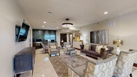Located at 2500 Brush Rd in Schaumburg, Illinois, this elegantly appointed single-floor amenity space blends upscale design with functional versatility. The main lounge area serves as the heart of the space, anchored by a tufted velvet sofa, patterned accent chairs, and a glass-top coffee table, all set atop a decorative area rug over gleaming large-format tile flooring. A striking crystal chandelier adds a refined focal point overhead, while curated large-scale artwork in gold, grey, and white tones lends a gallery-like ambiance throughout. Recessed lighting and floor lamps contribute to a warm, layered illumination scheme. Adjacent to the lounge, a dedicated business center features a built-in granite countertop workstation with multiple computer terminals, set against a dramatic textured mosaic tile accent wall that adds bold visual contrast. A separate conference room offers a rich mahogany-toned table surrounded by executive chairs, with natural light filtering through window treatments and geometric gold pendant light fixtures overhead, alongside a mounted display screen. A well-equipped kitchenette with dark wood cabinetry, granite countertops, a mosaic tile backsplash, and appliances provides a convenient refreshment area. Throughout the space, cohesive gold accents, sophisticated furnishings, and thoughtful design details create a polished, professional atmosphere.
