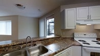 This single-story space includes a kitchen that serves as a functional and visually appealing area. The kitchen features granite-style countertops with warm brown and gold toning, providing a sturdy and attractive work surface that wraps around the space. A stainless steel sink with a gooseneck faucet is positioned along the counter, offering practicality and a clean aesthetic. White raised-panel cabinetry lines the upper walls, providing ample storage while contributing to a bright, classic look. A white appliance, likely a range, is partially visible to the right, integrating seamlessly with the overall color palette. Natural light filters into the kitchen through a window fitted with vertical blinds, offering a view of the surrounding outdoor area, including a parking lot and neighboring structures. The walls are painted in a neutral tone that complements the cabinetry and countertops, creating a cohesive and tidy environment. The ceiling is standard height and finished in white, with a visible HVAC vent contributing to the room's climate control. Overall, the space presents a clean, functional kitchen layout suited for everyday use.