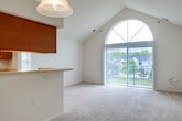 This single-floor residential unit offers a well-organized layout with a clean, neutral aesthetic throughout. The standout feature of the main living area is its dramatic vaulted ceiling, which rises to a striking arch and is paired with a large arched window fitted with vertical blinds. This design element draws in abundant natural light and provides views of the surrounding neighborhood and greenery, creating an open and airy atmosphere. The living space is finished with plush, light-toned wall-to-wall carpeting that adds warmth underfoot.

The galley-style kitchen is efficiently designed, featuring warm honey-toned wood cabinetry, light countertops, and tile flooring. It is equipped with a gas range, refrigerator, and an in-unit washer/dryer area tucked conveniently nearby. A pendant light fixture overhead adds a finishing touch to the kitchen's functional design.

The bedroom continues the neutral palette with soft carpeting and a large window dressed with vertical blinds, allowing for flexible light control. The full bathroom is well-appointed with warm wood vanity cabinetry, a single sink with a wide mirror, tile flooring in a terracotta tone, and a full tub and shower enclosure. The overall ambiance of the space is bright, clean, and move-in ready.
