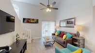 This two-story residence is defined by its dramatic vaulted ceilings in the main living area, which create a striking sense of openness and volume. Light-toned hardwood-style flooring runs throughout the lower level, complementing the bright, neutral walls and enhancing the airy atmosphere. A ceiling fan anchors the living room, where a sliding glass door fitted with vertical blinds invites natural light and provides access to an outdoor area. A staircase with white balusters leads to the upper level, which overlooks the living space through an open railing, reinforcing the home's open and connected feel.

The kitchen features rich, dark wood cabinetry, dark granite-style countertops, and black appliances, including a range, built-in microwave, and refrigerator. It flows naturally into a dedicated dining area with a pendant light fixture. Both bedrooms are located on the upper level, each offering plush carpeting, ample closet space, and access to natural light. One bedroom includes a sliding glass door leading to a private balcony. The two bathrooms are finished with dark countertops, white cabinetry, and tub/shower combinations dressed in coordinating striped curtains.