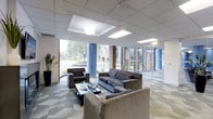 This single-floor lounge space presents a thoughtfully designed interior that blends contemporary style with comfortable, functional furnishings. The room is anchored by a striking feature wall adorned with vertical grey and silver striped wallpaper, adding depth and visual interest to the space. Recessed lighting throughout the ceiling provides consistent, ambient illumination across the entire area.

The main seating arrangement centers around a colorful grouping of armchairs in bold orange and yellow tones, positioned around a low coffee table and set atop a vibrant, patterned area rug. This creates an inviting focal point for conversation and relaxation. Two wall-mounted flat-screen televisions are positioned for easy viewing from multiple seating angles.

Towards the rear of the room, a high-top table with bar-style seating offers an alternative gathering spot. The flooring features a distinctive herringbone-patterned tile, adding a refined architectural element to the overall design. A hallway visible toward the back leads to additional areas, suggesting further amenities beyond the main lounge. The overall ambiance is energetic yet welcoming, with a modern, hospitality-inspired aesthetic.