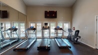 This fitness center is a comprehensive, single-floor facility designed to support a full range of workout routines. The space is divided into distinct functional zones, seamlessly connected to create an open and energizing environment.

The cardio area is anchored by a row of treadmills equipped with individual digital consoles, accompanied by stationary and recumbent bikes. A wall-mounted flat-screen television overlooks this section, while a bank of French doors and multi-pane windows floods the area with natural light and offers views of the surrounding outdoor greenery. Reflective wall mirrors expand the sense of space and allow users to monitor their form.

The strength training zone features a robust selection of equipment, including Cybex and cable-based multi-functional machines, a lat pulldown and cable crossover station, and a dedicated press machine. Vaulted ceilings rise dramatically in this section, contributing to an open, airy atmosphere. Full-length mirrors line the walls, further enhancing the spacious feel.

An additional strength area provides space for functional training and accessory work, complete with cable machines and open floor space. Durable rubber flooring throughout the entire facility ensures safety and easy maintenance. The facility is well-ventilated with recessed lighting and climate control, creating a comfortable training environment year-round.