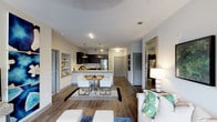 This two-bedroom, two-bathroom single-floor residence offers an open-concept design that connects the living, dining, and kitchen areas into one cohesive and functional space. Hardwood-style flooring runs throughout, lending warmth and continuity to the layout.

The kitchen is equipped with stainless steel appliances, including a refrigerator and microwave, and features dark cabinetry paired with light stone countertops. A kitchen island with seating provides an additional casual dining option. Pendant lighting hangs above the island, adding both task lighting and a stylish focal point. Built-in shelving adjacent to the kitchen offers additional display or storage space.

The dining area accommodates a small table and chairs, positioned between the kitchen and living room to create a natural flow for entertaining. The living room provides ample space for comfortable seating arrangements.

A hallway leads to the two bedrooms and bathrooms, each accessed through paneled doors. The neutral color palette of soft whites and grays throughout creates a clean, contemporary ambiance. Recessed lighting supplements the overall brightness of the space, contributing to a welcoming and well-lit interior.