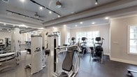 This single-level fitness center is thoughtfully designed to accommodate a comprehensive range of workout routines. The space is organized into distinct functional zones, each dedicated to a specific type of exercise. A cardio section lines one wall, housing a row of Cybex treadmills positioned beneath a large window that brings in natural light and views of surrounding greenery. Adjacent to this area, multiple elliptical trainers and upright cardio machines are arranged to maximize floor space while maintaining an open, uncluttered feel.

The strength training section is outfitted with an extensive selection of professional-grade Cybex cable and weight machines, as well as a tiered dumbbell rack with a flat bench for free weight exercises. Full-length mirrors along one wall visually expand the space and allow for proper form monitoring during workouts. French-style glass-paned doors connect the various zones, enhancing openness and flow throughout the facility.

Architecturally, the space features coffered ceilings with decorative beams, crown molding, and a combination of recessed and track lighting that ensures bright, even illumination. Dark rubber flooring provides both durability and safety underfoot, complementing the clean, neutral wall tones and lending the facility a polished, professional ambiance overall.