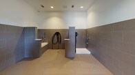 This pet-friendly amenity space combines an expansive outdoor dog park with a well-appointed indoor wash and grooming station. The outdoor area features a generously sized, gravel-covered ground surface enclosed by sturdy metal fencing, providing a secure environment for pets to roam freely. Several young trees are planted throughout the park, offering shade and greenery, while the surrounding open views add to the airy atmosphere. The park is situated adjacent to a natural stone and panel-clad building, with gated entry points for easy access.

The indoor dog wash facility is thoughtfully designed with functionality in mind. The interior features large-format tile flooring and wainscoting in a warm taupe tone, complemented by clean white upper walls and recessed lighting overhead. The main wash room is equipped with two dedicated grooming stations — one featuring a raised tub with a handheld sprayer and an adjacent wall-mounted pet dryer with a flexible hose, and a second walk-in style shower station. A glass-paneled door connects the wash room to the outdoor park, allowing seamless transitions between the two spaces. Additional open areas within the facility provide room for drying and grooming, complete with a floor drain and trash receptacle for convenient cleanup.