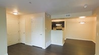 This single-floor residence features a clean, functional layout with warm neutral walls and dark hardwood-style flooring that flows throughout the main living area, creating a cohesive and modern aesthetic. The open-concept design connects the living space to a semi-open kitchen, which is defined by a half-wall pass-through that provides separation while maintaining visual connectivity. The kitchen is equipped with dark cabinetry on both the upper and lower sections, a built-in range with an overhead vent hood, and a microwave, offering a practical and stylish cooking area. Recessed HVAC vents are positioned throughout the ceiling and lower walls, ensuring efficient climate control. The living area benefits from multiple doorways, suggesting access to additional rooms or storage spaces, and white six-panel doors contribute to the crisp, clean finish of the interior trim work. White baseboards frame the perimeter of the space, complementing the neutral palette. The overall ambiance is bright, open, and move-in ready, with a straightforward layout designed for practical everyday living.