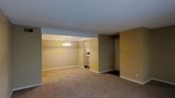 This single-floor space features an open-concept layout that seamlessly connects the living area, dining space, and kitchen. The living and dining areas share a continuous expanse of wall-to-wall carpeting in a neutral beige tone, creating a cohesive and comfortable feel underfoot. Warm tan and taupe-painted walls contribute a cozy, understated ambiance throughout.

A defining feature of the dining area is a classic multi-arm chandelier that serves as a focal point, providing both functional lighting and a touch of traditional elegance. The transition between the living and dining zones is defined by a subtle architectural soffit overhead, adding gentle structure to the open floor plan.

A doorway visible from the main living area leads into the kitchen, where white cabinetry and appliances are partially visible, suggesting a functional and clean layout. Near the kitchen, a standard entry door provides access to the unit, framed by neutral walls and clean white baseboards that run consistently throughout the space. The textured ceiling and straightforward design give the space a practical, move-in-ready character suitable for a variety of furnishing styles.