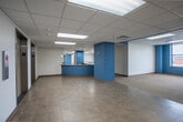 Located at 500 W Pershing Rd in Kansas City, Missouri, this single-floor commercial office space offers a well-organized and functional layout suited for a variety of professional uses. The space is accessed via a lobby area featuring two elevators framed by chrome trim, tiled flooring, and distinctive teal-painted structural columns that serve as a consistent design element throughout the floor.

The open office areas are expansive and carpeted in neutral tones, providing flexible configurations for workstations or collaborative zones. Bold teal accent walls and columns add visual character against the predominantly white walls and drop tile ceilings illuminated by recessed fluorescent panel lighting. Structural columns are thoughtfully integrated throughout, defining zones without fully enclosing them.

Private offices line the perimeter, several of which feature windows overlooking the surrounding Kansas City urban landscape, allowing natural light to filter into the space. Warm wood-trimmed doors with sidelight glass panels contribute a professional aesthetic throughout the corridors and office entries.

A dedicated kitchenette provides ample cabinetry, extended countertops, a sink, and a dishwasher, making it well-equipped for daily staff use. Long corridors connect the various sections of the floor, creating a logical and navigable flow. Overall, the space presents a clean, ready-to-occupy commercial environment with practical amenities and a cohesive design identity.