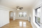This single-story space showcases a well-maintained interior defined by classic architectural details and a clean, neutral palette. The room pictured features warm-toned hardwood floors that extend throughout, lending a sense of continuity and warmth to the space. Crown molding frames the white ceiling, adding a refined, traditional character, while a ceiling fan with an integrated light fixture serves as both a functional and decorative focal point overhead. Natural light filters in through a window fitted with blinds, brightening the room and highlighting the pristine white walls. A built-in closet with paneled doors provides generous storage, while an elegant arched doorway leads to an adjoining area, hinting at thoughtful architectural design throughout. A window air conditioning unit offers climate control. The overall ambiance is light, airy, and move-in ready, combining practical features with charming details that give the space a comfortable and inviting character suited to a variety of uses.