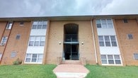 This single-floor unit is situated within a well-established brick building, identified as Building D. The exterior showcases a traditional tan brick facade with a distinctive arched entryway, wall-mounted lighting, and a central staircase that services multiple levels. Large windows line the front of the building, allowing for generous natural light throughout. A neatly maintained grassy lawn leads to a concrete walkway at the entrance.

Inside, the unit is accessed through a solid white entry door equipped with both a knob lock and deadbolt for added security. The living area is comfortably sized, featuring wall-to-wall carpeting in warm neutral tones and a large window fitted with sheer curtains that diffuse natural light throughout the space, creating a bright and welcoming atmosphere. The neutral-toned walls provide a versatile backdrop throughout the unit.

Storage is addressed through a dedicated closet fitted with wire shelving racks, offering practical organization space. Tile flooring is visible within the closet area, providing a durable surface. The overall interior maintains a clean, functional aesthetic with white trim accents and brass hardware details. Residents also have access to an outdoor grilling area, adding a convenient amenity to the property.