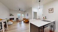 This single-floor residence showcases a thoughtfully designed open-concept layout that seamlessly connects the kitchen and living area. The kitchen is a standout feature, equipped with dark cabinetry, stainless steel appliances, and a white quartz peninsula that doubles as a breakfast bar with barstool seating. A pendant light with a glass cylinder shade adds a contemporary touch above the peninsula, which also houses a sink and dishwasher. Light wood-look flooring runs throughout the main living areas, contributing to a bright, cohesive aesthetic.

The living room is comfortable and well-appointed, featuring a wall-mounted fireplace, a ceiling fan, a wall-mounted mini-split HVAC unit, and a glass door that opens to the outdoors, allowing natural light to fill the space. The bedroom offers plush carpeting, a large window with outdoor views, and a relaxed, cozy atmosphere. The bathroom includes dark wood cabinetry, a single-sink vanity, and a shower with a decorative curtain. A dedicated laundry room with full-size washer and dryer units and overhead shelving adds practical convenience. The overall ambiance is modern, clean, and welcoming throughout.
