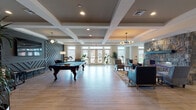 Located at 400 Boulder Pass in Oxford, CT, this single-story community clubhouse is thoughtfully designed to serve as a premier gathering and recreational hub. The main lounge area is expansive and open, anchored by a coffered ceiling with recessed lighting and elegant pendant fixtures. Light wood-look flooring runs throughout, enhancing the bright, welcoming atmosphere. A full-size billiards table anchors the game area, while a stone-faced fireplace with a sleek mantle serves as the focal point of the sitting area, furnished with teal sofas and woven accent chairs. Large windows and glass-paneled double doors flood the space with natural light and provide outdoor views. A dedicated lounge nook features a bold geometric accent wall in deep gray, complemented by a sectional sofa and tray ceiling detail. The fully equipped kitchen boasts a contemporary design with warm wood cabinetry, glossy white upper cabinets, white quartz countertops, subway tile backsplash, stainless steel appliances, and a large island with bar seating — all illuminated by under-cabinet lighting and pendant fixtures. A glass-walled studio space houses a grand piano and an adjacent mirrored fitness or dance room, offering versatile programming options. The overall ambiance is modern, polished, and inviting.