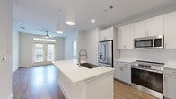 This thoughtfully designed two-bedroom, two-bathroom residence on Nexus Ave in Stafford, TX offers a clean, contemporary aesthetic across a single floor. The open-concept kitchen and living area forms the heart of the unit, anchored by a large white quartz center island with an undermount sink and pendant lighting overhead. The kitchen is equipped with stainless steel appliances, including a French-door refrigerator, slide-in range, and built-in microwave, complemented by white upper cabinetry, gray lower cabinets, and a subtle tile backsplash. Light wood-style flooring runs throughout the main living areas, which benefit from natural light through French doors topped with a transom window and a ceiling fan above. Both bedrooms feature neutral carpet and windows that bring in additional daylight. Each bathroom is finished with white quartz vanities, LED-backlit mirrors, and wood-look flooring, with access to a dedicated closet featuring built-in shelving. The overall palette of white, gray, and warm wood tones creates a bright, cohesive, and modern living environment.