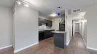 This single-story residence offers a well-organized, functional layout designed for comfortable everyday living. The kitchen serves as a central highlight, featuring dark espresso-toned cabinetry, light granite-style countertops, a double-basin stainless steel sink, a peninsula with additional counter space, and black appliances including a refrigerator and dishwasher. A striking blue pendant light and track lighting add a contemporary touch to the kitchen area.

Wood-look flooring runs consistently throughout the unit, providing a warm, cohesive aesthetic. The open entryway leads into a spacious living area with neutral gray walls and clean white baseboards, creating a bright and airy atmosphere. Track lighting near the entry enhances the welcoming feel.

The bedrooms are generously sized and feature ceiling fans with integrated lighting. One bedroom showcases a distinctive painted brick accent wall alongside a window with blinds that brings in natural light. The rooms offer ample space for a variety of furniture arrangements.

The bathrooms are clean and functional. One features a full tub-shower combo with white subway tile surround, while the second includes a tub-shower unit with wood-look flooring and a dark-toned vanity. Both bathrooms maintain a crisp, minimal aesthetic consistent with the rest of the unit.