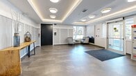 Located at 2699 Peatt Rd in Langford, BC, this building lobby offers a polished and welcoming first impression with a thoughtfully designed interior. The space is finished with large-format stone-look tile flooring that lends a clean, sophisticated aesthetic throughout. A distinctive coffered ceiling with integrated ambient LED strip lighting and flush-mount ceiling fixtures creates a layered lighting scheme that enhances the sense of depth and warmth.

The lobby is divided into functional zones. Near the entrance, a full-width entry mat greets residents and guests, accompanied by a bank of individual mailboxes and parcel lockers — providing convenient mail and package management. A rustic live-edge console table serves as a focal point along one wall, adding a natural contrast to the contemporary surroundings.

A comfortable seating area features two tufted barrel chairs with blue accent cushions arranged around a round nesting side table, positioned beside a large window that allows natural light to filter in and offers views of the surrounding greenery. A large-scale abstract mural anchors the adjacent wall, adding an artistic element to the space.

Wainscoting panels line the walls, contributing a refined architectural detail, while a secure access door provides controlled entry to the building's interior corridors. The overall ambiance is modern, clean, and inviting.