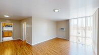 This single-floor unit presents a clean and functional layout with a warm, neutral aesthetic throughout. The open living area is finished with rich hardwood-style flooring that flows seamlessly across the main spaces, complemented by smooth white ceilings with flush-mount lighting. Large windows dressed with vertical blinds allow generous natural light to fill the rooms, creating a bright and airy atmosphere. A wall-mounted air conditioning unit provides climate control in the main living space. The kitchen is equipped with granite countertops, warm honey-toned cabinetry, a double-basin sink, and an over-the-range microwave, offering both practicality and style. The bathroom features a dark espresso vanity topped with a granite countertop, a large mirror with overhead lighting, a sliding glass shower enclosure, and light tile flooring — providing a tidy and well-finished space. A separate bedroom benefits from its own large window with vertical blinds, continuing the hardwood-style flooring and maintaining the unit's cohesive, understated design. The overall ambiance is modest yet well-maintained, making this a practical and comfortable space.