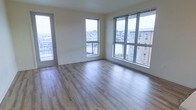 This single-floor residence offers a clean, contemporary design characterized by an open and airy atmosphere. The main living area is finished with smooth light-toned hardwood flooring that extends throughout, lending warmth and continuity to the space. Large floor-to-ceiling windows line the exterior walls, flooding the interior with an abundance of natural light and providing expansive views of the neighboring residential buildings and landscaped grounds below. A glass-panel balcony door opens directly from the living area, seamlessly connecting the interior to the outdoor space and enhancing the sense of openness. The neutral, light-toned walls serve as a versatile backdrop, complementing the natural finishes throughout. The generous proportions of the living area allow for flexible furniture arrangements, accommodating a variety of lifestyle needs. The overall ambiance is modern and welcoming, with the interplay of natural light, clean lines, and quality finishes creating a comfortable and functional living environment. The views of the surrounding community visible through the multiple windows add a connected yet private feel to the space.