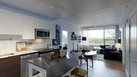 This single-floor space showcases a thoughtfully designed open-concept layout that seamlessly connects the kitchen, dining, and living areas into one cohesive living environment. The kitchen is outfitted with stainless steel appliances, including a range and over-the-range microwave, complemented by crisp white cabinetry and a subway tile backsplash. A substantial island with a stainless steel countertop serves as both a functional workspace and a casual gathering point, complete with seating.

The dining area transitions naturally from the kitchen, featuring a round table and mid-century modern chairs that add warmth to the otherwise contemporary palette. The living area is anchored by large floor-to-ceiling windows dressed with sheer curtains, flooding the space with natural light and offering elevated views of the surrounding tree canopy and skyline beyond.

Track lighting runs along the smooth ceiling, providing flexible illumination throughout. The interior design balances cool tones — whites, grays, and navy — with warm wood and metallic accents. The overall ambiance is modern and urban, with an airy, well-lit atmosphere.