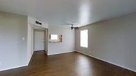 This single-story space presents a clean, straightforward layout designed for practical living. The main living area is anchored by rich, dark hardwood flooring that extends throughout, offering a warm contrast against the neutral white walls and crisp white ceiling. A ceiling fan is centrally mounted, providing both air circulation and a functional focal point overhead.

Natural light enters through two windows — one positioned along the side wall and another illuminating the entryway area — keeping the space feeling open and airy. The front entry features a solid white door with a clean, simple frame, providing a defined and secure point of access.

A pass-through window connects the living area to the kitchen, offering a convenient open communication between the two spaces. Warm wood cabinetry is visible through the pass-through, and a sink is positioned just below it, hinting at a compact but efficiently arranged kitchen beyond.

The living area itself is generously proportioned, allowing for flexible furniture arrangements. Crisp white baseboards frame the flooring throughout, adding a finished, polished detail. The overall ambiance is neutral, minimal, and move-in ready, suited to a variety of interior styles.