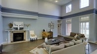 This single-floor space makes a striking impression through its living area, which boasts soaring double-height ceilings that create an expansive, airy atmosphere. The room is defined by a bold two-tone wall treatment, with a cool blue-grey upper register separated from the lower walls by a prominent decorative crown moulding detail, adding architectural character throughout.

The living area is anchored by a classic fireplace with a white mantel surround, accompanied by built-in storage and a display shelf above. Elevated clerestory windows, including a distinctive diamond-paned transom, allow natural light to pour in from above, while additional side windows offer views to the exterior brick surroundings.

Dark hardwood flooring runs throughout, grounding the space with warmth and contrast against the cool wall palette. Recessed lighting complements the natural light, ensuring the room remains bright and welcoming. The overall layout accommodates generous seating arrangements, and the combination of traditional architectural details with contemporary furnishings gives the space a refined yet approachable ambiance.