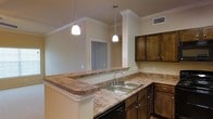 This single-floor residence centers around a thoughtfully designed kitchen that serves as the heart of the space. The kitchen features rich, dark-stained wood cabinetry that provides generous storage, complemented by granite-look laminate countertops that wrap around the perimeter and extend into a peninsula-style breakfast bar. This raised bar counter creates a natural division between the kitchen and the adjacent living area, making it ideal for casual dining or entertaining.

A double-basin stainless steel sink is positioned along the main counter run, paired with a chrome faucet. Overhead, two pendant lights with frosted white shades hang at varying heights, casting warm, focused light across the workspace. Crown molding lines the upper edges of the walls and cabinets, adding a refined architectural detail throughout.

The kitchen opens visually into a connected living or dining area, visible through the pass-through design of the peninsula. Neutral warm-toned walls and light flooring enhance the sense of openness throughout. A ceiling vent is positioned for efficient climate control, and the overall layout reflects a practical, open-concept design suited to everyday living.