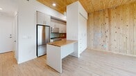 Located at 29 Crown Street in New Haven, CT, this thoughtfully designed single-floor studio offers an efficient and modern living experience with distinctive architectural character. The space is defined by its striking exposed mass timber ceilings and natural wood-paneled walls, which lend warmth and texture throughout. Light-toned hardwood flooring complements the wood elements and ties the space together cohesively.

The combined kitchen and living area forms the heart of the unit. The kitchen is well-appointed with white cabinetry, stainless steel appliances including a full-size refrigerator and gas range, warm butcher block countertops, and a peninsula that doubles as a dining or workspace. Pendant lighting adds a contemporary accent above the peninsula.

The living area benefits from a large picture window that frames views of the New Haven cityscape, flooding the space with natural light and providing an urban backdrop. The exposed wood walls in this area create a cozy, cabin-like atmosphere contrasted by the modern finishes throughout.

A clean, all-white bathroom includes a full soaking tub with shower, toilet, and vanity with wood-look tile flooring. An in-unit stacked washer and dryer offers added convenience, alongside a dedicated closet for storage. The entrance provides a welcoming transition into this compact yet complete urban retreat.