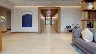 This single-floor leasing office and lobby presents a polished, upscale environment designed to make a strong first impression. The entrance greets visitors with floor-to-ceiling glass double doors that flood the space with natural light, framed by lush tropical greenery and decorative sculptural accents on elegant console tables. Large-format light stone tile flooring flows seamlessly throughout, lending a clean and cohesive foundation to the entire space.

The reception area features a sleek, modern desk clad in warm wood tones and crisp white surfaces, accented by striking brass pendant light fixtures overhead and tall wall sconces that add a warm, ambient glow. Textured stone-look wall panels lend depth and character to the surroundings, while curated abstract artwork — including a bold blue canvas — adds artistic personality.

A wide corridor with warm wood-paneled walls connects the lobby to additional areas, maintaining a refined and purposeful design language throughout. The lounge area is a standout, featuring two generously proportioned gray sofas facing each other across a substantial wood coffee table, all anchored by a dramatic abstract blue-and-gray area rug. Built-in wood shelving with curated décor and ample recessed lighting complete the sophisticated atmosphere. Tropical plants are thoughtfully placed throughout, adding organic warmth and vibrancy to this elegantly appointed space.