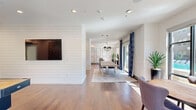 Located at 12639 Coit Rd in Dallas, TX, this thoughtfully designed single-floor community space offers a generous and versatile layout across four distinct areas. Warm hardwood flooring flows seamlessly throughout, anchoring a refined aesthetic that blends modern and transitional design elements.

The main gathering area showcases elegant wainscoting and decorative wall paneling, recessed lighting, and a tufted velvet sofa arrangement centered around a coffee table — ideal for relaxed socializing. Bold gold artwork adds a striking focal point against the crisp white walls.

An open dining area features a substantial wood-and-chrome dining table beneath a statement chandelier, set against a backdrop of floor-to-ceiling sliding glass doors and bold navy patterned drapes. These doors open to views of a resort-style pool, bathing the space in abundant natural light.

A secondary seating nook with a bistro-style table and chairs provides a more intimate setting, also benefiting from direct pool views through large windows. Tall tropical plants throughout add a lush, inviting ambiance.

The fully equipped kitchen is a standout feature, lined with shiplap-clad walls, white shaker cabinetry, quartz countertops, a marble tile backsplash, and stainless steel appliances including a refrigerator, double wall oven, and built-in microwave — perfectly suited for catering and community events.