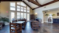 This single-story clubhouse combines rustic charm with functional design. The main gathering area is defined by dramatic vaulted ceilings adorned with dark-stained exposed wood beams, creating a striking architectural focal point. A sweeping wall of floor-to-ceiling windows floods the interior with natural light while offering views of the surrounding outdoor environment, enhancing the open, airy atmosphere of the space.

Dark slate tile flooring grounds the room and complements the warm tones of the solid wood furniture throughout. Multiple rustic wooden tables paired with matching chairs provide flexible seating arrangements, suitable for both casual gatherings and organized activities. A dedicated workstation area along one wall is equipped with a computer and printer, offering residents a convenient business center within the clubhouse.

Additional storage is provided by a large wooden armoire, consistent with the space's rustic aesthetic. Wall sconces contribute warm accent lighting, while a connected hallway leads to additional areas of the facility. The overall ambiance balances a welcoming, lodge-inspired aesthetic with practical community amenities.