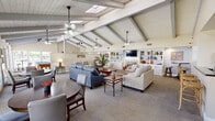 Located at 1401 S Harbor Blvd in La Habra, California, this single-floor space offers a generously sized, open layout designed for comfortable gathering and recreation. The living area is immediately distinguished by its dramatic vaulted ceilings with exposed painted wood beams and white tongue-and-groove planking, complemented by multiple skylights and ceiling fans that fill the space with natural light and airflow. Multiple seating arrangements are spread across the expansive carpeted floor, creating distinct zones for conversation and relaxation. A tiled fireplace serves as a focal point at one end of the room, flanked by a built-in white cabinetry unit with open shelving and a wall-mounted flat-screen television above. Additional built-in shelving units provide display and storage space throughout. A pool table situated near a wall of windows adds a recreational dimension, with views of the surrounding greenery enhancing the bright, open atmosphere. Track lighting and pendant fixtures provide layered illumination throughout. The kitchen features warm wood cabinetry, dark granite countertops, tile flooring, and stainless steel appliances including a refrigerator, range, and microwave — offering full functionality for catering and events. Overall, the space presents a welcoming, versatile environment suited for community events, social gatherings, and everyday recreation.