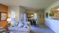 This single-floor residence showcases an open-concept design that integrates the living and dining areas into one cohesive, flowing space. The living area is anchored by a plush, neutral-toned sofa accented with colorful decorative pillows, creating a comfortable and inviting atmosphere. Warm beige walls and soft carpeting throughout contribute to the overall warmth of the interior.

The dining area sits adjacent to the living space and features a dark wood dining table with matching chairs, illuminated by a pendant light that adds a focused, intimate ambiance to the space. A framed piece of wall art and decorative metal wall accents add visual interest to the room.

Natural light filters in through a sliding glass door dressed with patterned curtains, offering access to an outdoor area and brightening the interior. An arched doorway adds an architectural detail that enhances the open flow between spaces. The vaulted ceiling creates a generous sense of vertical space, making the overall layout feel expansive and airy. The design balances functionality with comfort, making it well-suited for both relaxed everyday living and casual entertaining.