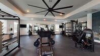 This spacious fitness center is thoughtfully designed to support a full range of workout routines. The main exercise area features durable rubber flooring throughout, providing both safety and comfort during training sessions. A striking oversized ceiling fan with multiple blades is mounted within a tray ceiling, complemented by recessed lighting that ensures the space is bright and well-illuminated.

The strength training section is comprehensively outfitted with a multi-station cable and pulley machine, a power rack with weight plates, a leg press/hack squat machine, and a full dumbbell rack — all reflected in large wall-mounted mirrors that visually expand the space. Flat-panel TVs are mounted on the walls for entertainment during workouts.

The cardio zone includes multiple treadmills, stationary bikes, and elliptical machines, each equipped with individual display screens. Natural light filters in through several windows lining the perimeter, offering views of the surrounding outdoor area and creating an open, airy atmosphere.

Near the entrance, a dedicated alcove finished with deep blue-grey tile houses a dual-basin stainless steel water fountain with a bottle-filling station. A glass-panel door provides direct outdoor access, connecting the fitness center conveniently to the exterior.