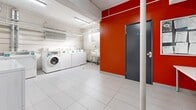Located at 550 Eglinton Ave W in Toronto, this shared laundry facility offers a well-organized and functional space for building residents. The room is finished with light grey tile flooring throughout and features white-painted walls accented by a bold orange feature wall near the entrance, adding a splash of color to the utilitarian space. The laundry area is equipped with a row of top-load washing machines and front-load washers, complemented by a pair of dryers, all arranged along the perimeter walls for efficient use of the floor space. A stainless steel utility sink with exposed plumbing is positioned along one wall, providing a convenient handwashing or pre-treatment station. An electronic payment kiosk is mounted near the machines, enabling cashless transactions. Overhead, exposed conduit, pipes, and ventilation ducts run along the ceiling, reflecting the functional, no-frills design of the space. A small frosted window allows a measure of natural light into the laundry area. Adjacent to the main laundry zone is a separate waiting area, furnished with a white table and informational notices posted on the wall, along with a fire extinguisher and waste bin. Dual grey steel doors provide access to and from the facility, ensuring a secure and accessible environment for all users.