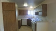 This single-floor space centers around a practical, efficiently designed kitchen that makes good use of its available footprint. The kitchen features warm-toned wooden cabinetry along both the upper and lower walls, providing generous storage throughout. A white gas range with multiple burners anchors one side of the kitchen, accompanied by an overhead range hood for ventilation. A white dishwasher is seamlessly integrated beneath the counter on the opposite side, adjacent to a double-basin sink fitted with a standard faucet. The countertops feature a light-colored laminate surface that contrasts cleanly with the darker cabinetry. A window fitted with vertical blinds above the sink area allows natural light to filter into the space, brightening the work area. Two flush-mounted ceiling light fixtures provide consistent overhead illumination throughout the kitchen. The flooring appears to be a neutral-toned vinyl or tile, suited for easy maintenance in a high-use area. The textured ceiling adds a subtle architectural detail consistent with the overall utilitarian design of the space. The layout follows a U-shaped configuration, maximizing workflow efficiency between the cooking, cleaning, and preparation zones.