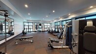 Located at 311 El Camino Real in San Carlos, California, this single-level fitness center is thoughtfully designed to support a comprehensive workout experience. The space is generously proportioned, with a layout that accommodates a wide range of exercise equipment without feeling crowded. Recessed ceiling lighting is distributed evenly throughout, complemented by an array of large, dark-framed windows along multiple walls that invite natural light and offer views of the surrounding outdoor environment, including glimpses of a pool area.

The cardio zone features a row of treadmills and elliptical machines positioned along the windowed walls, each equipped with individual display screens. Strength training is well-represented with cable machines, a multi-functional weight station, adjustable benches, and a rowing machine. A dedicated functional training area includes a heavy punching bag and a medicine ball rack. A large wall-mounted mirror on one side of the room enhances the sense of space and supports form-conscious training.

Durable rubber flooring in a tile pattern covers the entire workout area, providing traction and impact absorption. The entry vestibule features olive-toned wainscoting, tile flooring, a water bottle filling station, and a keypad-secured glass door, adding both convenience and a polished aesthetic to the overall facility.