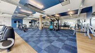Located at 222 East Boyd Drive in Baton Rouge, Louisiana, this expansive 4,860-square-foot fitness center is a well-appointed, single-floor facility designed to support a wide range of workout routines. The space is organized across nine distinct zones, offering a comprehensive exercise environment with a modern, clean aesthetic throughout.

The main strength training area features hardwood flooring and floor-to-ceiling mirrored walls, a full dumbbell rack, weight plates, benches, and a bold "Stay Strong" wall mural. Exposed industrial-style ductwork runs across the ceiling, complementing the contemporary design. Adjacent to this is a large open workout floor finished in blue modular carpet tiles, lined with cable machines, selectorized strength equipment, and flat-screen TVs mounted to blue accent walls.

A dedicated cardio zone benefits from expansive floor-to-ceiling windows that flood the space with natural light and provide outdoor views of the surrounding pool and terrace area. Treadmills, ellipticals, stationary bikes, and rowing machines are arranged for optimal use of this bright, airy section.

A flexible studio space features light hardwood floors, textured accent walls, wall-mounted TVs, and room for group fitness or personal training. Additional rooms provide storage and accessory equipment, including medicine balls and fitness mats. Stairwells with steel railings and tile flooring connect the facility's circulation areas, completing this thoroughly equipped fitness destination.