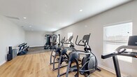 Located at 110 Peach Blossom Rd in Warner Robins, Georgia, this dedicated fitness center offers a thoughtfully designed exercise space within a single, open-plan room. The interior is finished with neutral gray walls and recessed lighting that keeps the environment bright and welcoming. The flooring transitions between light-toned wood-look planks and durable rubber gym matting, effectively delineating the cardio zone from the strength training area.

The cardio section is well-stocked with multiple elliptical trainers and treadmills, providing ample options for aerobic workouts. A multi-station cable machine serves as the centerpiece of the strength training zone, which is complemented by a tiered dumbbell rack holding a comprehensive range of free weights. A stationary bike rounds out the equipment selection, supporting a variety of low-impact training routines.

Natural light enters through several windows fitted with blinds, while a glass-panel exit door at one end of the room provides additional brightness and convenient outdoor access. Recessed ceiling fixtures ensure consistent illumination throughout the space. A water station is also visible near the exit, adding a practical amenity for users. The overall layout is efficient and uncluttered, making this fitness center a functional and user-friendly facility.