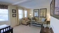 This single-floor residence offers a warm and inviting atmosphere, thoughtfully designed with comfort and classic style in mind. The living area serves as the heart of the home, bathed in natural light streaming through multiple large windows dressed with elegant patterned curtains. The neutral color palette — featuring soft beige walls, plush carpeting, and coordinating upholstered furnishings — creates a calm, cohesive ambiance throughout the space. A sectional sofa and a matching armchair are arranged around a sleek glass-top coffee table, providing a comfortable and functional seating arrangement ideal for relaxing or entertaining. Decorative framed botanical prints adorn the walls, adding a refined, traditional touch to the room. A classic table lamp provides warm accent lighting, complementing the natural light and enhancing the overall coziness of the space. Exposed ceiling beams add a subtle architectural detail that gives the room character and depth. The layout flows naturally, making efficient use of the available space while maintaining an open and uncluttered feel. Overall, this residence combines timeless design elements with practical comfort, creating a welcoming environment that is both functional and aesthetically pleasing.