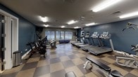This fitness center is thoughtfully laid out across a single floor, offering distinct zones to accommodate a variety of workout styles. The main exercise area features a checkered rubber floor and is lined with an extensive array of cardio equipment, including multiple treadmills, ellipticals, stationary bikes, and a cycling bike. Overhead ceiling fans and fluorescent lighting keep the space comfortable and well-lit, while windows along one wall provide natural light and outdoor views. A large wall mirror enhances the sense of space and supports strength and form training. An adjacent studio room offers a quieter setting for stretching, cycling, or mat-based workouts, finished with a speckled cork-style floor, recessed lighting, sage-green walls, and a full-length framed mirror. A yellow bench and yoga equipment are also present in this area. The entry corridor connects the two workout zones and features tile flooring, blue upholstered seating, colorful wall art, and access to restroom facilities. An exterior door in the main workout room provides convenient direct access to the outdoors. Overall, the fitness center presents a clean, functional, and inviting environment suited for daily exercise.
