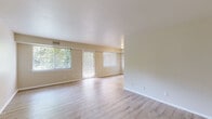 This single-floor residence offers a cohesive, move-in-ready layout finished with light wood-style flooring that runs throughout the entire unit, creating a clean and consistent aesthetic. The open living area is generously proportioned, featuring neutral beige walls, a sliding glass door, and large windows that invite abundant natural light and views of surrounding greenery. The space is bright and airy, lending itself well to a variety of furniture arrangements. The kitchen is equipped with light oak cabinetry, dark granite-style countertops, a stainless steel sink, and a black gas range with overhead ventilation — a practical setup for everyday cooking. Multiple bedrooms are present, each finished with the same wood-style flooring, neutral walls, and windows fitted with horizontal blinds for privacy and light control. The rooms are well-sized and versatile. The unit includes two bathrooms: one featuring a standalone glass-enclosed shower stall alongside a separate soaking tub and vanity, and a second with a full shower-tub combo and vanity. Both bathrooms are finished in clean white tones with light flooring. Overall, the space presents a tidy, functional design with consistent finishes and ample natural light throughout.