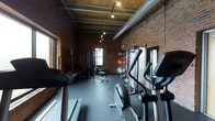 This fitness space combines industrial design with functional amenities, creating a unique workout environment. Exposed red brick walls and wooden ceiling beams lend the space a raw, warehouse-like aesthetic, while large windows allow natural light to filter in. The high ceilings, supported by visible ductwork and structural beams, contribute to a spacious, open feel throughout the room.

The flooring consists of durable dark rubber matting, well-suited for heavy equipment and high-impact exercise. Track lighting mounted along the ceiling provides focused illumination across the space. The equipment layout is thoughtfully organized, with cardio machines—including treadmills, ellipticals, and a stationary bike—positioned along one side, while cable machines and strength training stations occupy the opposite area. Stability and medicine balls are also available, catering to a variety of workout routines. The combination of classic industrial architecture with modern fitness equipment gives this space a distinctive character that balances form and function effectively.