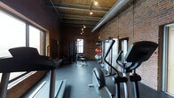 This fitness space combines industrial design with functional amenities, creating a unique workout environment. Exposed red brick walls and wooden ceiling beams lend the space a raw, warehouse-like aesthetic, while large windows allow natural light to filter in. The high ceilings, supported by visible ductwork and structural beams, contribute to a spacious, open feel throughout the room.

The flooring consists of durable dark rubber matting, well-suited for heavy equipment and high-impact exercise. Track lighting mounted along the ceiling provides focused illumination across the space. The equipment layout is thoughtfully organized, with cardio machines—including treadmills, ellipticals, and a stationary bike—positioned along one side, while cable machines and strength training stations occupy the opposite area. Stability and medicine balls are also available, catering to a variety of workout routines. The combination of classic industrial architecture with modern fitness equipment gives this space a distinctive character that balances form and function effectively.
