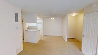 This single-floor residential space presents a clean, functional layout centered around an open-concept living and kitchen area. The living room is finished with wall-to-wall beige carpeting that adds warmth underfoot, while the textured white ceilings and crisp white walls create a bright, neutral backdrop throughout. A ceiling fan with a light kit is centrally positioned in the living area, providing both air circulation and ambient lighting.

The kitchen is connected to the living space via a convenient pass-through counter, which also serves as a casual bar-style seating ledge. White cabinetry lines the upper portion of the kitchen, offering ample storage, while the overall layout keeps the space feeling open and accessible. The pass-through design enhances the sense of connectivity between cooking and living areas.

An entry door is visible to the right, framed by white walls and a surface-mounted ceiling light, offering a welcoming transition into the space. Additional doorways suggest further rooms beyond the main living area. The overall aesthetic is simple and understated, with a consistent use of white tones and neutral flooring that makes the space feel clean, airy, and move-in ready. The layout is well-suited for everyday living, with a practical flow between functional zones.
