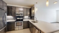 This single-floor residence offers a well-designed open-concept layout that seamlessly connects the kitchen and living areas, creating a bright and functional living environment. The kitchen is a standout feature, showcasing rich dark-toned shaker-style cabinetry paired with a classic subway tile backsplash and sleek light quartz countertops. A full suite of stainless steel appliances — including a refrigerator, electric range, built-in microwave, and dishwasher — provides a polished, contemporary finish. Pendant lighting hangs above the countertop, adding warmth and definition to the space. Light wood-style flooring runs throughout the main living areas, lending an airy, cohesive feel that extends into the open living room, which benefits from a ceiling fan and a large window allowing generous natural light to fill the room. The bedroom continues the bright aesthetic with plush neutral carpeting, a ceiling fan, and a sizable window with exterior views. The bathroom is finished with a wide dual-sink vanity topped with light quartz, a large framed mirror, and a tub-shower combination. The overall design balances modern finishes with a clean, neutral palette, creating a comfortable and inviting atmosphere throughout.