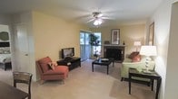 This single-floor residence offers a welcoming and functional layout centered around a comfortable living area. The main living room is a highlight of the space, featuring a classic fireplace with a stone surround and decorative mantel that serves as a natural focal point. Soft, neutral-toned carpet runs underfoot, contributing to the warm and inviting ambiance throughout. A ceiling fan with integrated lighting is centrally mounted, ensuring year-round comfort and even air circulation. Natural light flows generously into the room through a large sliding glass door framed by vertical blinds, offering views of lush greenery just outside. The space accommodates a well-arranged seating area with ample room for lounging and entertaining. Warm yellow walls lend a cheerful, bright character to the interior. A corner lamp and framed wall art add subtle decorative touches that enhance the overall aesthetic. The layout feels open and practical, with enough space to comfortably host both a media setup and a conversational seating arrangement. Overall, this single-floor home blends everyday functionality with a cozy residential charm, making it a well-rounded and livable space.