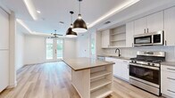 Located in Austin's vibrant 78704 zip code, this modern single-floor residence offers a thoughtfully designed open-concept layout with high-end finishes throughout. Light oak hardwood floors flow seamlessly across the main living areas, complementing the clean, neutral palette that defines the space.

The kitchen serves as the centerpiece of the home, anchored by a substantial island with a quartz countertop and built-in shelving. White and light wood cabinetry lines the walls, paired with stainless steel appliances including a gas range, built-in microwave, and French door refrigerator. A tray ceiling with integrated ambient lighting and bold black pendant fixtures adds architectural definition overhead. Open shelving and a built-in desk area enhance functionality.

The living area features a tray ceiling with cove lighting, a modern sputnik-style light fixture, and French doors that open to a private balcony with lush tree views, flooding the space with natural light.

The bedroom is generously proportioned with a large picture window framing greenery, a ceiling fan, and a dedicated walk-in closet with adjustable shelving systems. The primary bathroom is elegantly appointed with a walk-in glass shower featuring floor-to-ceiling tile surround, a dual-sink vanity with white cabinetry, and neutral tile flooring.