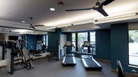 This fitness room offers a focused and functional workout environment designed to accommodate a range of exercise needs. The space is defined by deep navy blue walls that lend a modern, energetic atmosphere, complemented by a dark ceiling fitted with recessed lighting, linear LED fixtures, and multiple ceiling fans that ensure consistent air circulation throughout. The flooring features durable rubber gym matting, practical for both safety and equipment stability. Two treadmills are prominently positioned toward the front of the room, facing a row of windows that overlook an outdoor street scene, allowing natural light to filter in and providing a pleasant view during cardio sessions. Along one wall, a large mirror extends the perceived depth of the space while supporting strength and form-focused training. Additional equipment, including a weight bench and various strength training accessories, is arranged along the mirrored wall, offering versatility for resistance workouts. The windows on multiple walls enhance the sense of openness and bring in ample daylight. Overall, the room strikes a balance between compact efficiency and a well-considered layout, making it a practical and inviting space for daily fitness activities.